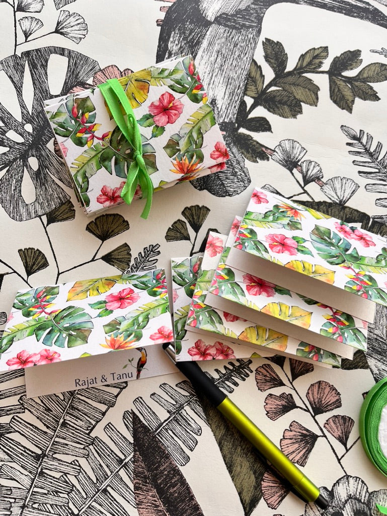 A styled flat-lay of tropical floral gift boxes and matching wrapped packages on a black-and-white botanical patterned surface. The boxes feature pink hibiscus and red flowers with green leaves, tied with bright green ribbon.