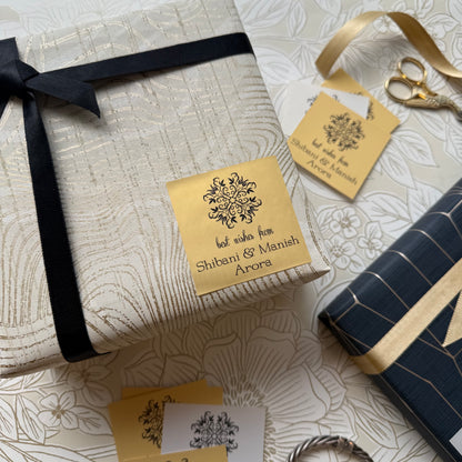 A styled flat-lay of gift wrapping supplies on a floral cream background, featuring a wrapped rectangular box with a black ribbon and a gold paper label, a navy wrapped box with gold stripes, scissors, and matching gold gift tags.