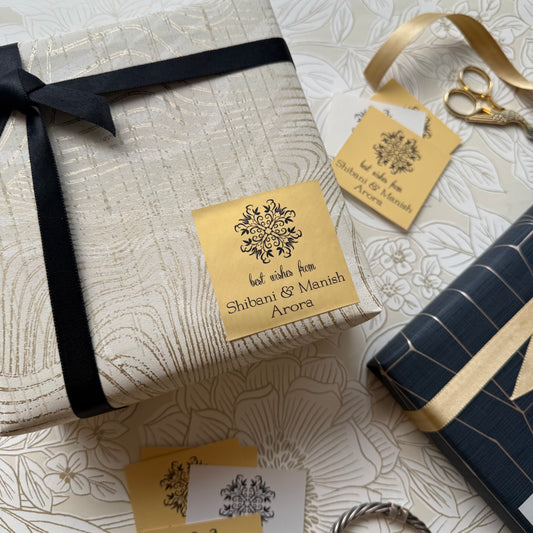 A styled flat-lay of gift wrapping supplies on a floral cream background, featuring a wrapped rectangular box with a black ribbon and a gold paper label, a navy wrapped box with gold stripes, scissors, and matching gold gift tags.