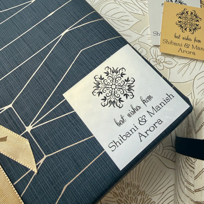 A navy blue fabric gift box or envelope with a white paper label that reads 'best wishes from Shibani & Manish Arora' and a black ornamental floral design. The box is placed on a beige patterned surface with a small gold card partially visible in the top right.