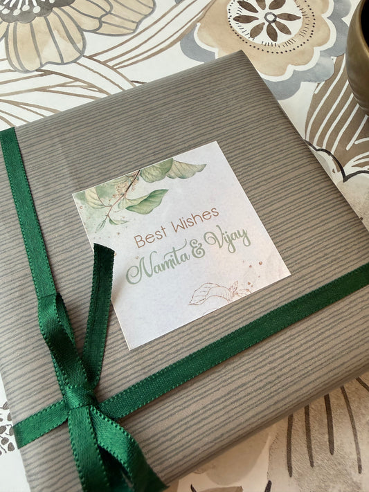 A square gift box wrapped in textured gray paper  and a personalised label that reads 'Best Wishes Namta & Vijay' with a leaf illustration, placed on a patterned surface.