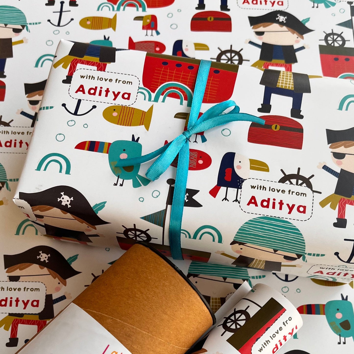 Nautical-themed wrapping paper with a pirate illustration pattern, tied with a turquoise ribbon and a kraft paper envelope, on a matching paper background. The text 'with love from Aditya' is printed on the wrapping paper.