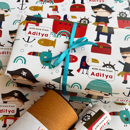 Nautical-themed wrapping paper with a pirate illustration pattern, tied with a turquoise ribbon and a kraft paper envelope, on a matching paper background. The text 'with love from Aditya' is printed on the wrapping paper.