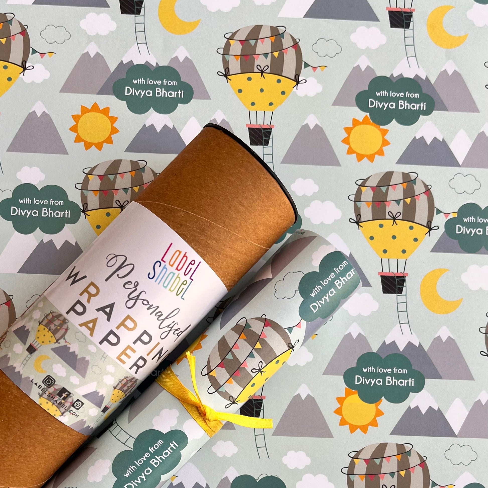 A roll of wrapping paper with a whimsical hot air balloon pattern, shown partially unrolled over a matching patterned surface. The wrapping paper features a light gray background with repeating illustrations of yellow crescent moons, brown and yellow hot air balloons, gray mountains, and green labels reading 'with love Divya Bharti'.