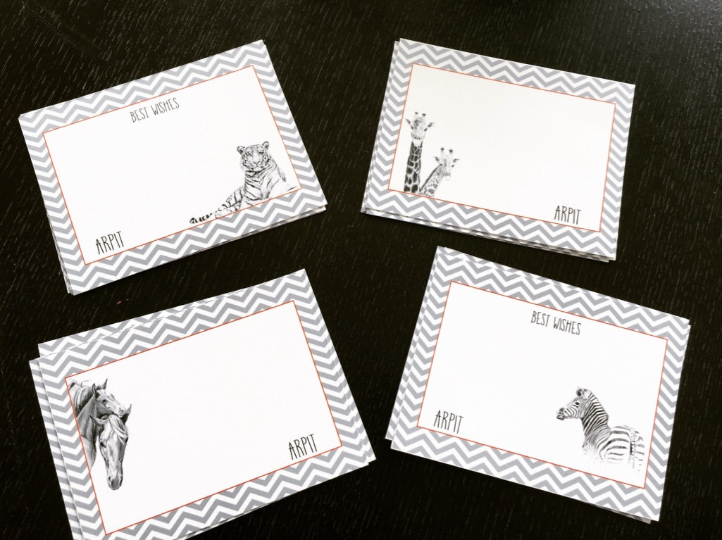 Four rectangular greeting cards arranged on a dark wooden surface. Each card has a gray and white chevron border, a white center, and a black-and-white animal illustration (tiger, giraffe, horse, zebra).