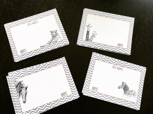 Four rectangular greeting cards arranged on a dark wooden surface. Each card has a gray and white chevron border, a white center, and a black-and-white animal illustration (tiger, giraffe, horse, zebra).