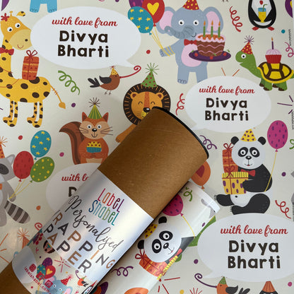 A personalized cardboard tube gift wrapper rolled up and tied with a red ribbon, placed on top of colorful birthday-themed wrapping paper printed with cartoon animals and the text 'with love from Divya Bharti'.
