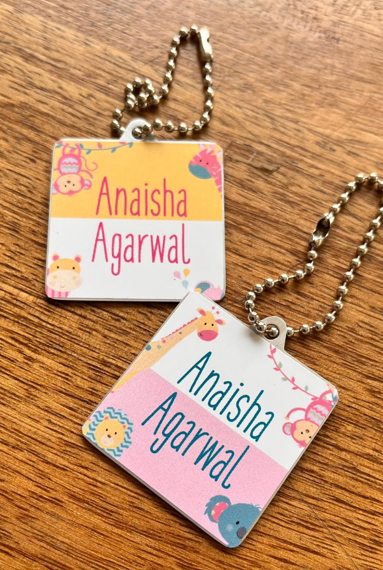 Two square metal keychains with ball chains, personalized with the text 'Anaisha Agarwal' and decorated with cute cartoon animal illustrations, resting on a wooden surface.