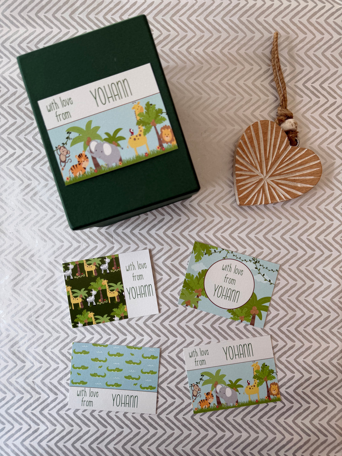 A set of personalized animal-themed cards and a dark green gift box with a colorful safari illustration, arranged on a gray and white chevron-patterned fabric next to a wooden heart ornament.