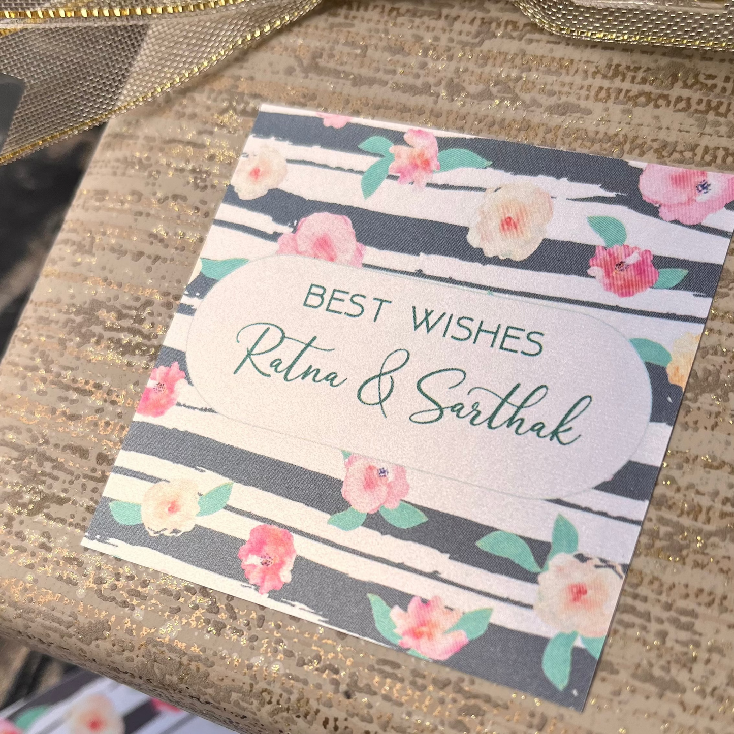 A close-up of a wrapped gift box tied with a gold ribbon, featuring a printed gift tag that reads 'BEST WISHES Ratna & Sarthak' with a floral design. Additional matching tags are visible on the surface below.