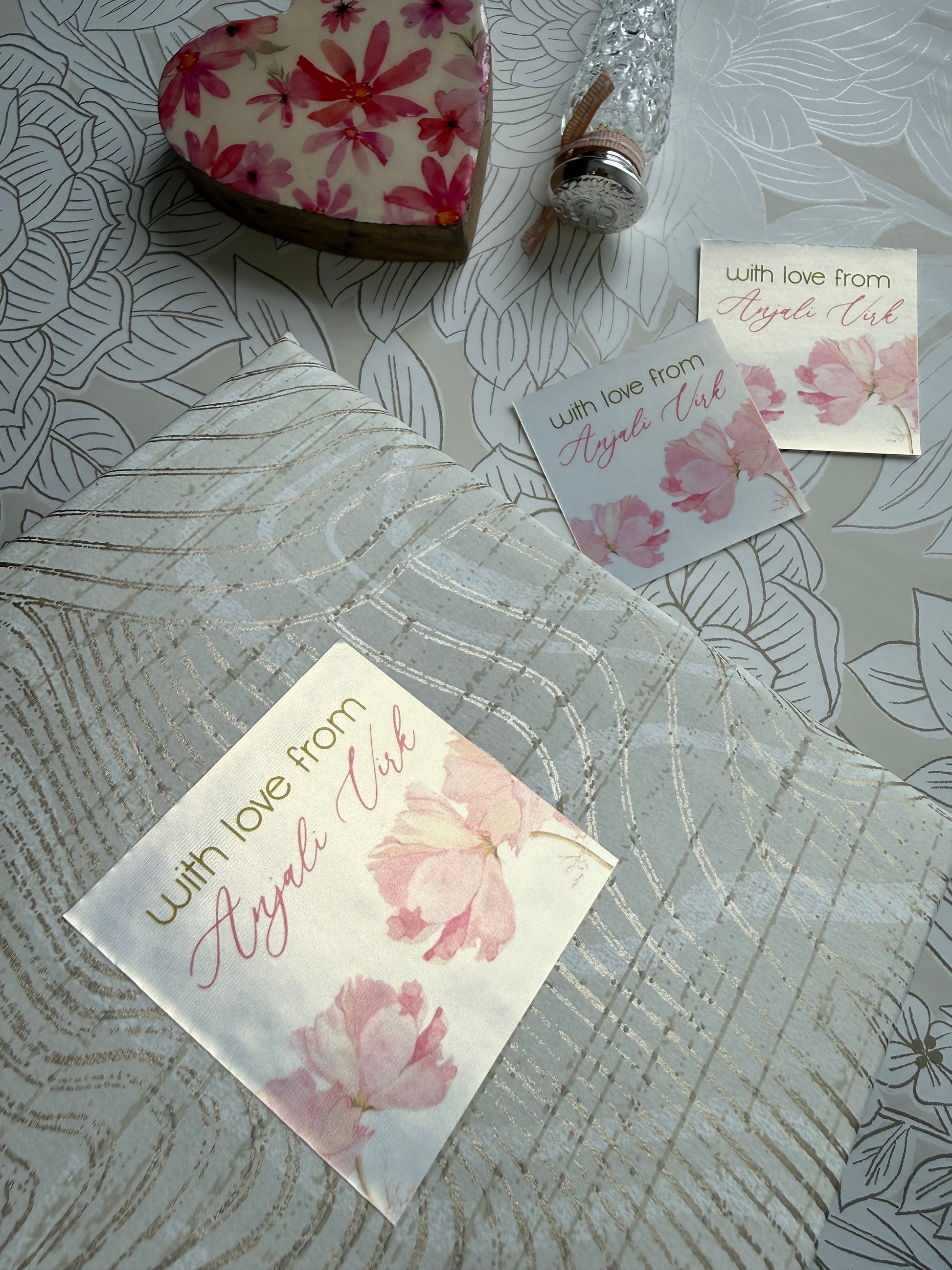 A styled flat-lay of a white and pink floral gift box with a clear patterned lid, accompanied by two matching floral cards and a decorative heart-shaped soap on a gray patterned background.