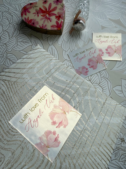 A styled flat-lay of a white and pink floral gift box with a clear patterned lid, accompanied by two matching floral cards and a decorative heart-shaped soap on a gray patterned background.