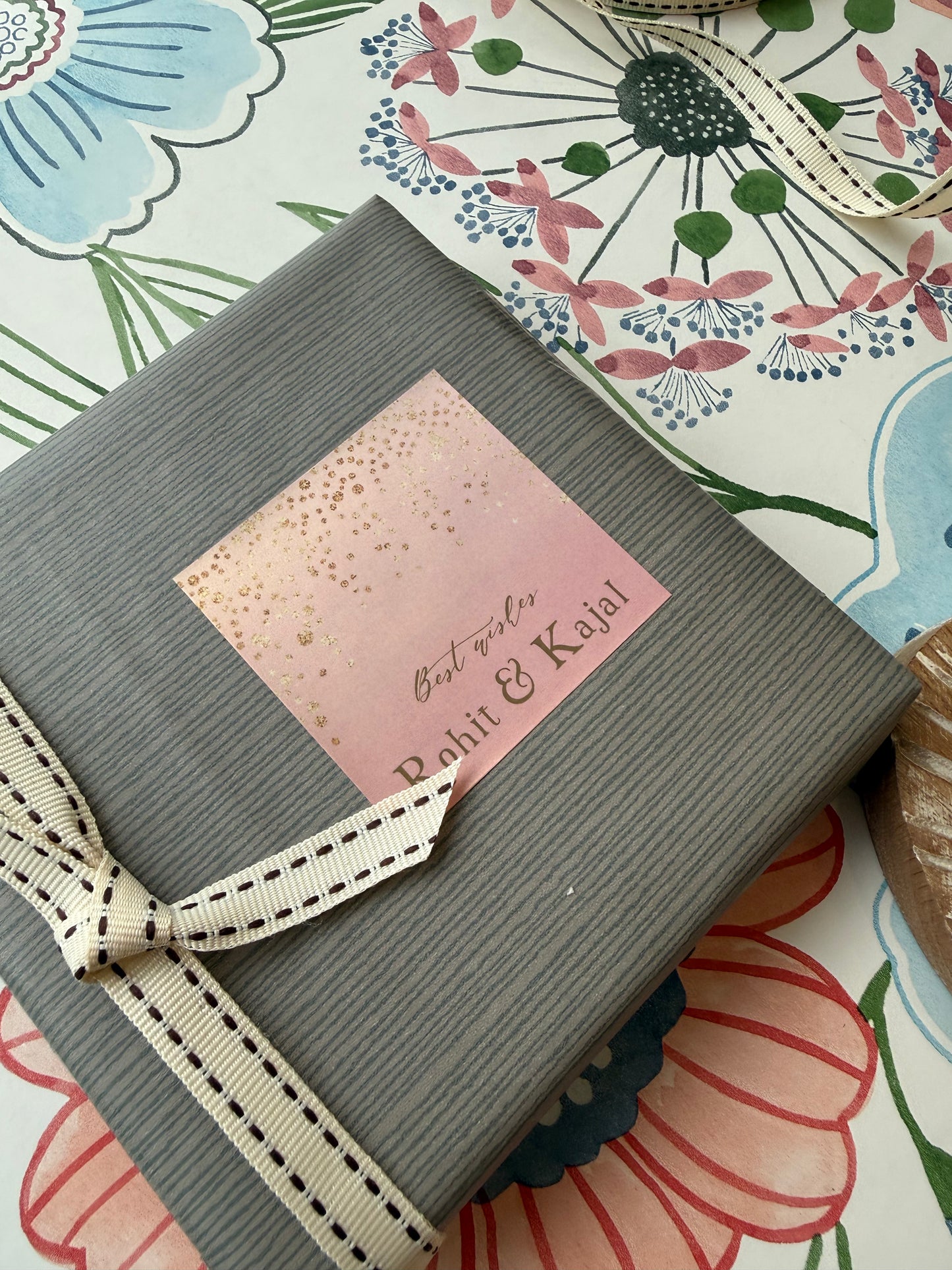 A gray ribbed gift box tied with a cream ribbon, featuring a pink square label that reads 'Best wishes Rohit & Kajal' in gold foil, placed on a floral-patterned surface.