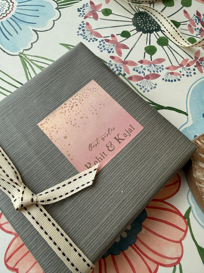 A gray ribbed gift box tied with a cream ribbon, featuring a pink square label that reads 'Best wishes Rohit & Kajal' in gold foil, placed on a floral-patterned surface.