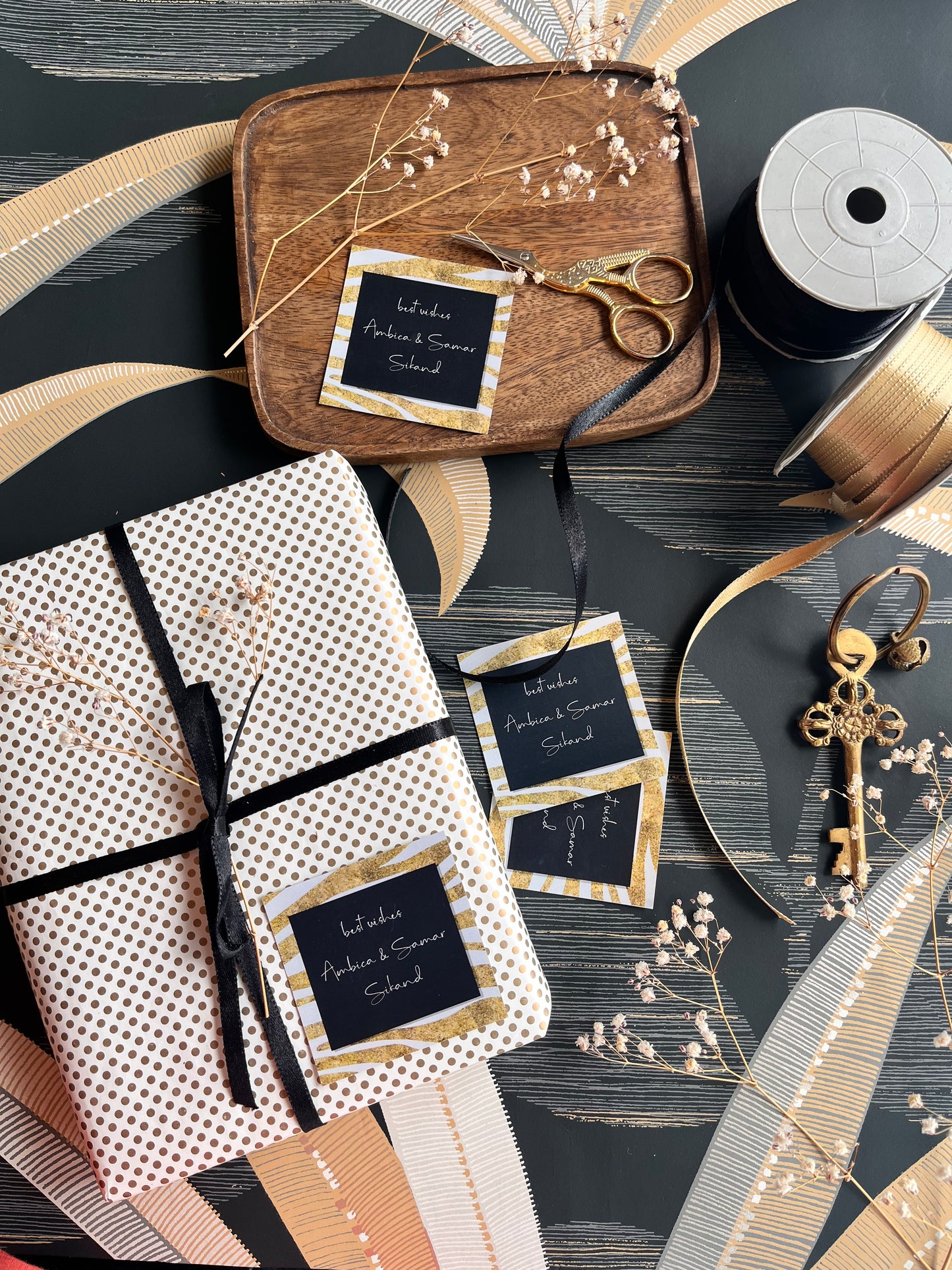 A styled flat-lay of gift wrapping supplies and a wrapped package on a dark wood surface. The main product shown is a gift box wrapped in white paper with small gold polka dots, tied with a black ribbon, and decorated with a black and gold tag.