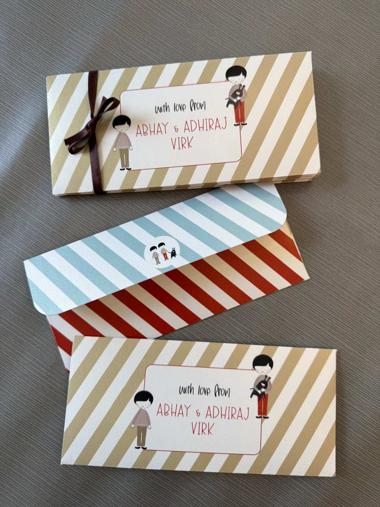 Three personalized gift boxes with striped patterns are arranged diagonally on a gray textured surface. The top box has beige and white diagonal stripes with a dark brown ribbon and a white label reading 'with love from ABHAY & ADHIRAJ VIRK'.