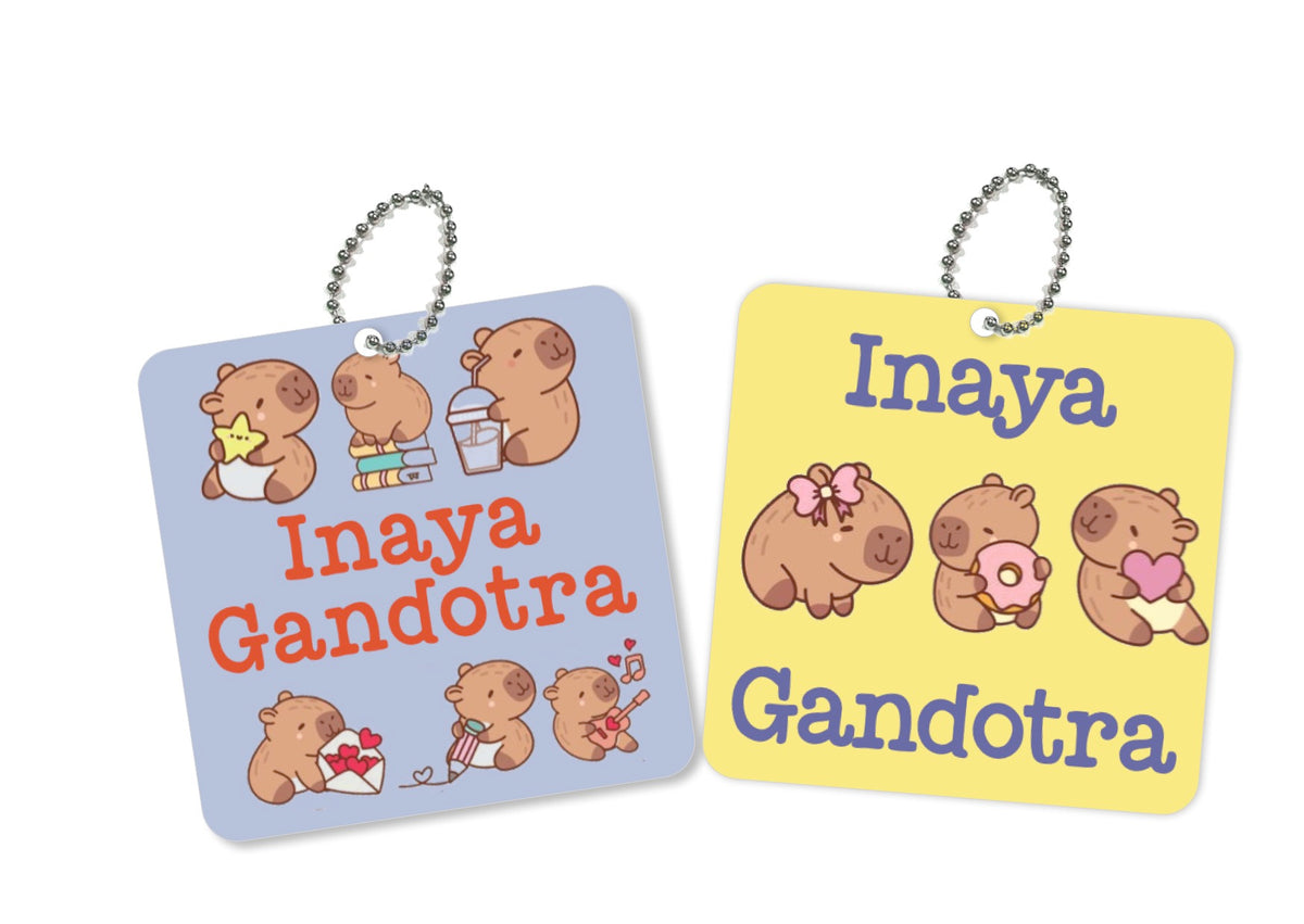 Two square metal  tags with rounded corners on a white background. One tag is light blue with red text reading 'Inaya Gandotra' and cute capybara illustrations.