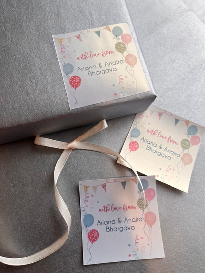 Three personalized gift tags with a colorful balloon and bunting design, attached to a gray wrapped gift box with a cream ribbon.