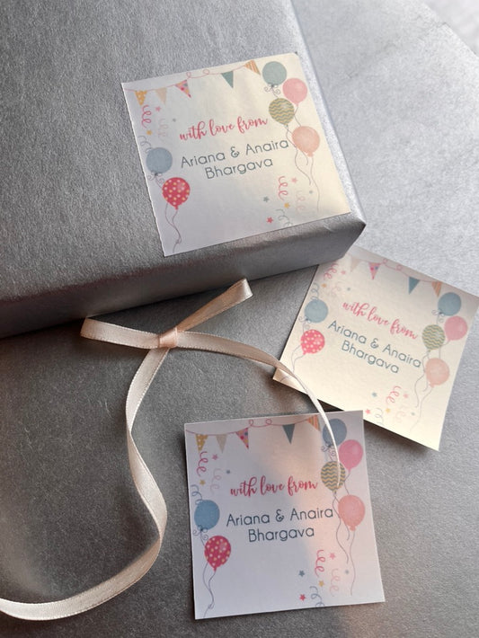 Three personalized gift tags with a colorful balloon and bunting design, attached to a gray wrapped gift box with a cream ribbon.