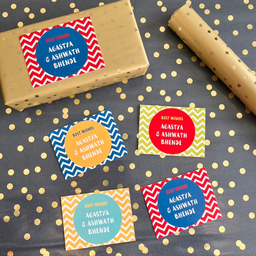 4 colorful gift wrapcardswith chevron and circular patterns are arranged on a dark gray background with gold polka dots. One label is shown wrapped around a gold gift box, and a roll of gold wrapping paper is visible in the upper right.