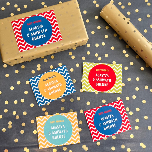 4 colorful gift wrapcardswith chevron and circular patterns are arranged on a dark gray background with gold polka dots. One label is shown wrapped around a gold gift box, and a roll of gold wrapping paper is visible in the upper right.