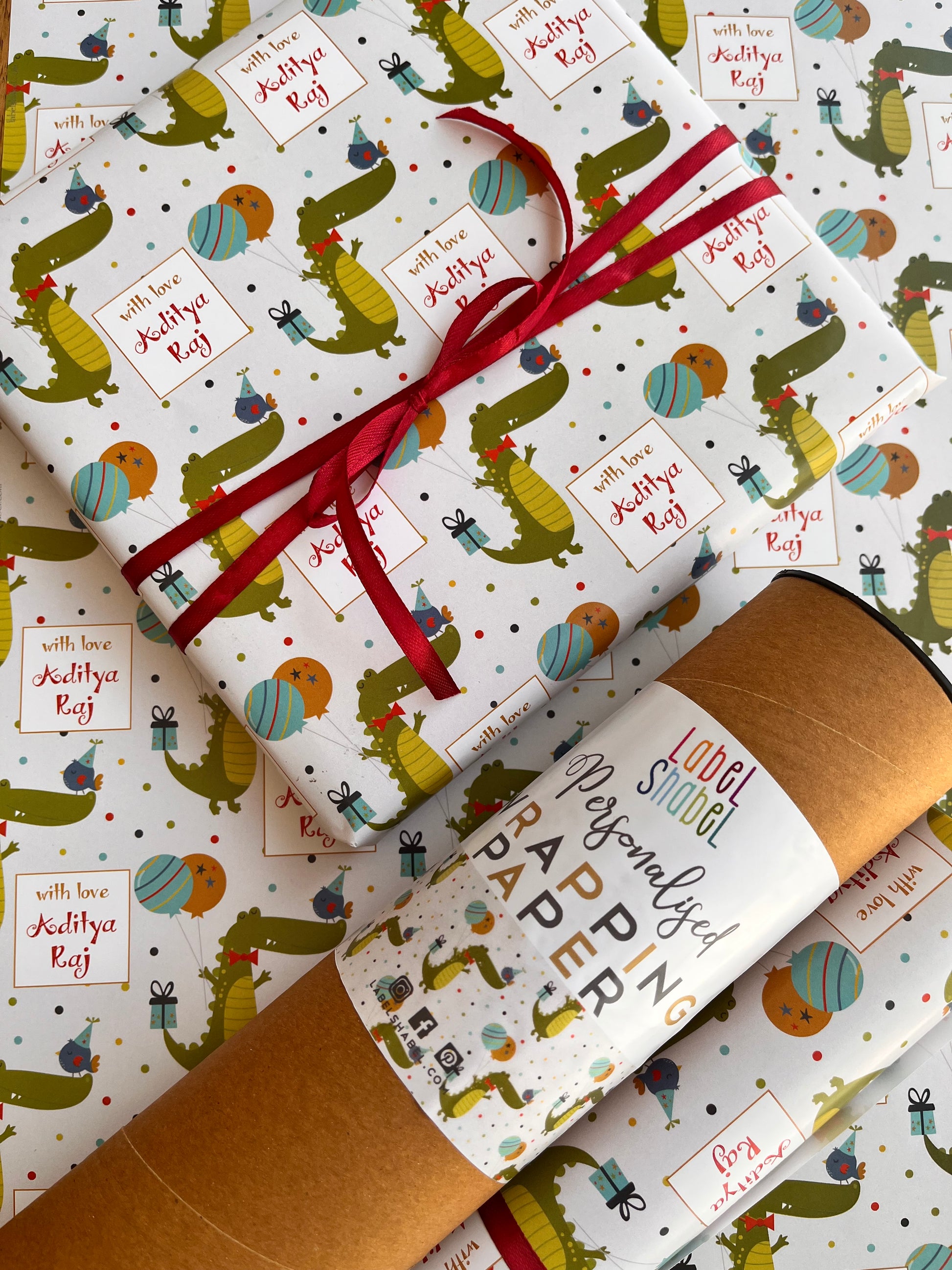 A roll of wrapping paper and a wrapped gift box with matching paper. The wrapping paper has a white background with a repeating pattern of green crocodiles carrying baloons and a gift