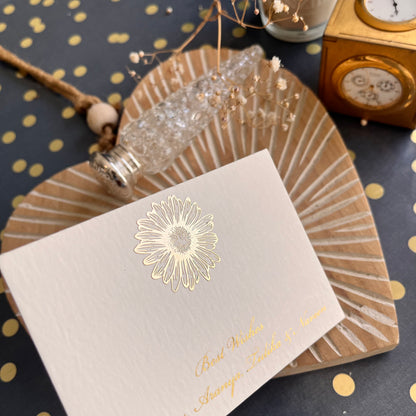 A flat card with a gold embossed daisy motif and matching lettering, displayed on a dotted background with a decorative element on the side.