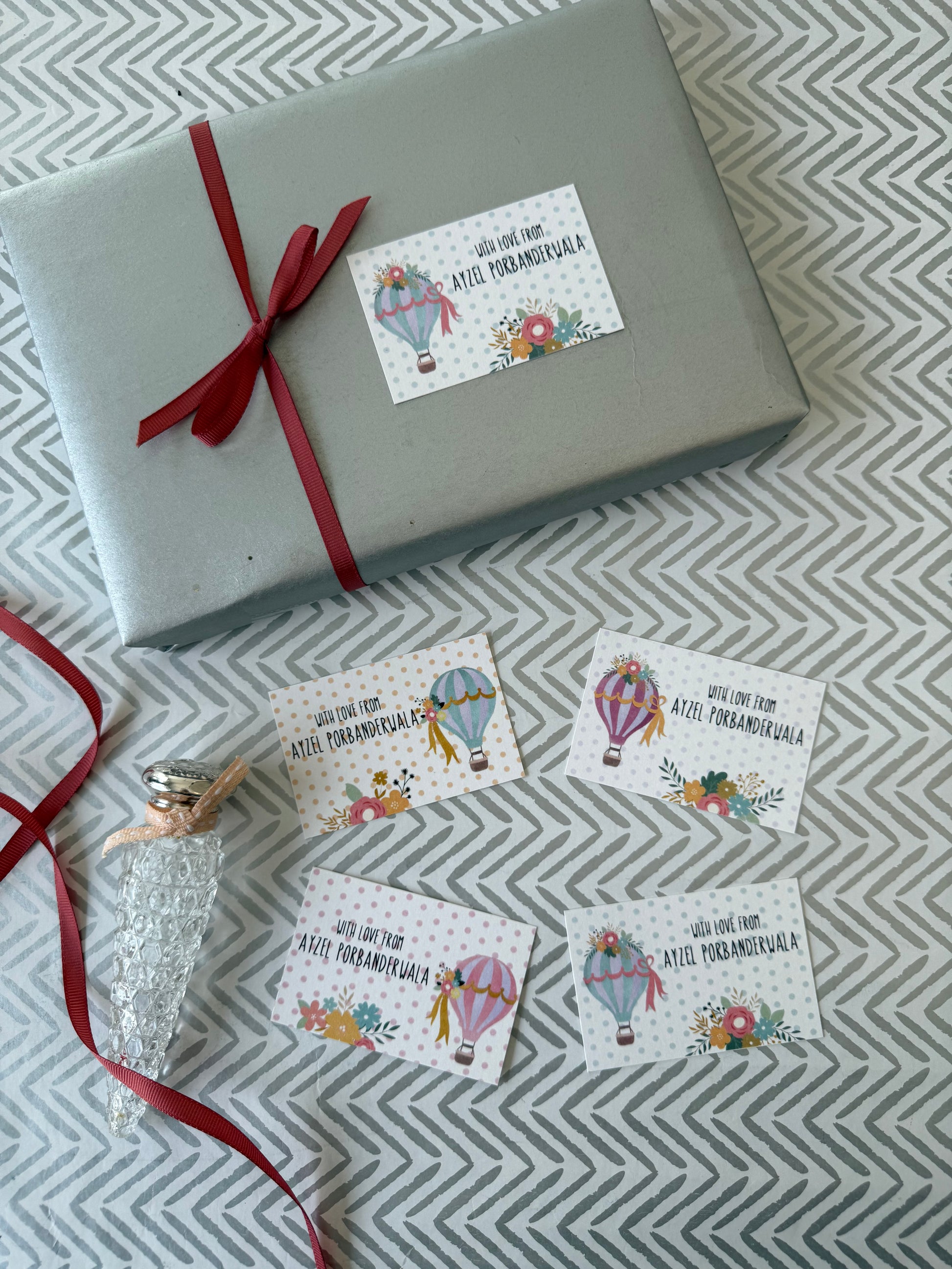 A wrapped gift box in light gray with a red ribbon and several small printed gift tags featuring hot air balloon illustrations and the text 'Will one day give to you Ate Pambankula' are arranged on a gray-and-white chevron fabric background.