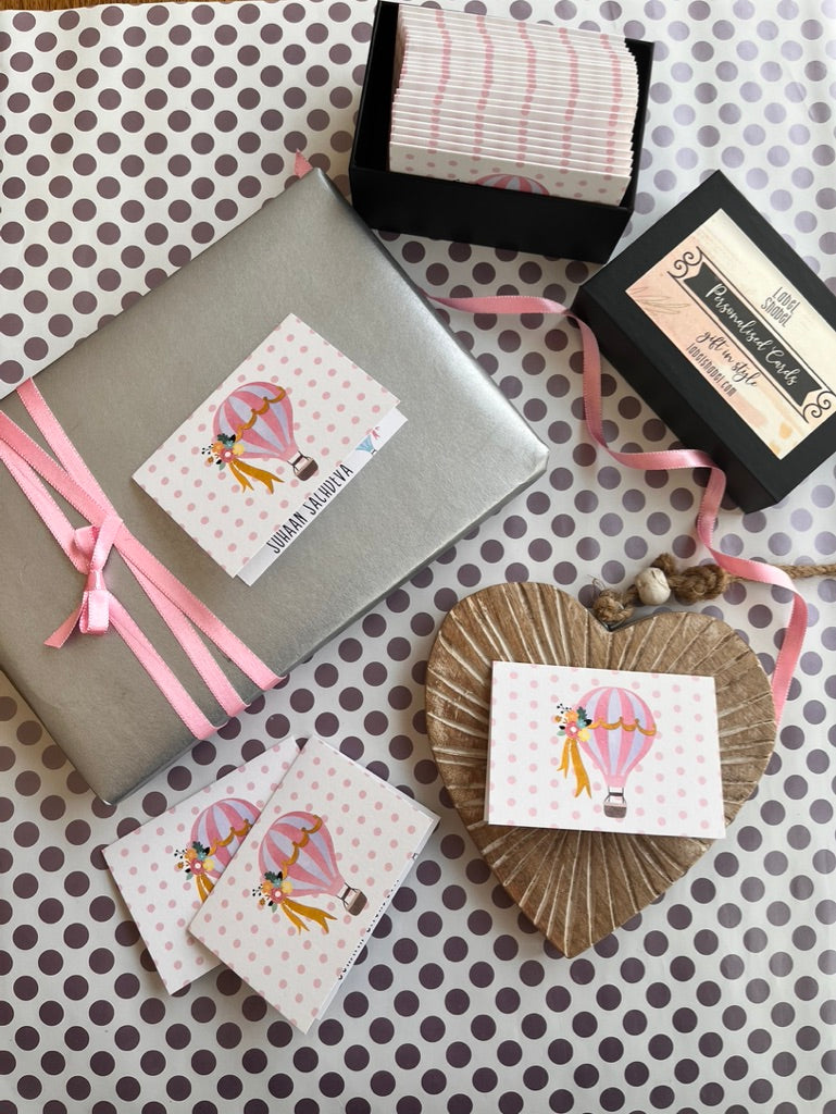 A styled flat-lay of gift-wrapping supplies and decorative paper products on a white backgroundfeautring a personalised gift tag with a hot air balloon illustration