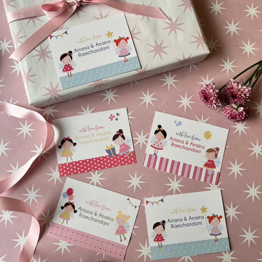 A styled flat-lay of a wrapped gift box and four matching printed cards on a pink star-patterned background. The gift is wrapped in white paper with a pale pink ribbon, and the cards feature cartoon girl illustrations, pastel decorative bands, and personalized text reading 'with love from Ariana & Anora Ramchandani'.