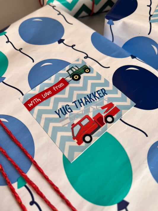 A close-up of a gift-wrapped package featuring a white paper wrap printed with large blue and teal balloons, a red ribbon, and a square decorative label that reads 'with love from yuG THAKKER' with toy vehicles on a blue chevron background.
