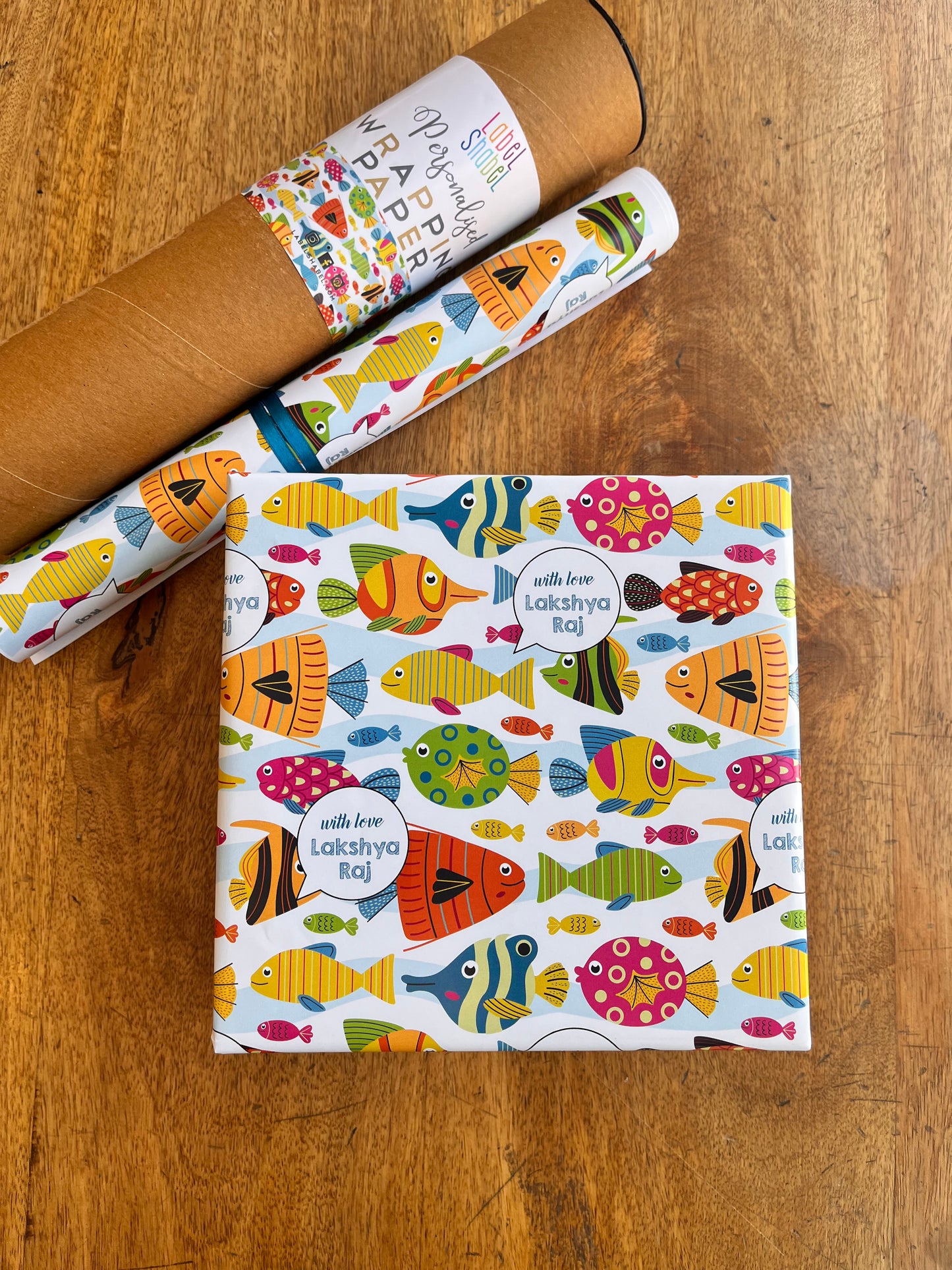 A roll of wrapping paper and a wrapped gift box on a wooden tabletop. The wrapping paper has a white background with a colorful pattern of fish