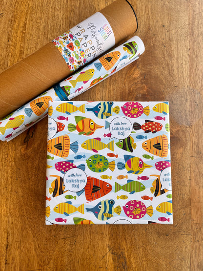 A roll of wrapping paper and a wrapped gift box on a wooden tabletop. The wrapping paper has a white background with a colorful pattern of fish