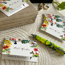Folded gift tag in white background with tropical leaves and birds and best wishes sign with name inside on top of a gift and a pen