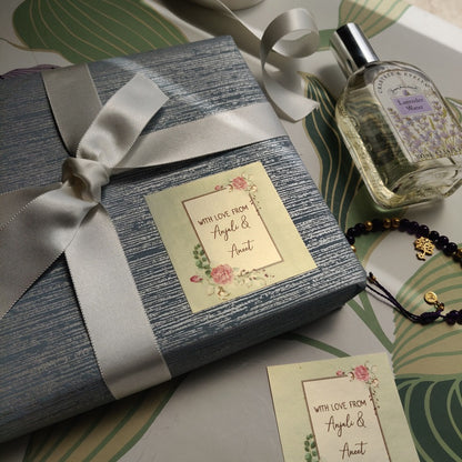 A wrapped gift box with a gray-blue textured wrap and a wide white satin ribbon, tied with a bow. A matching cream card and perfume bottle are arranged on a green tropical leaf-patterned surface next to a beaded bracelet.