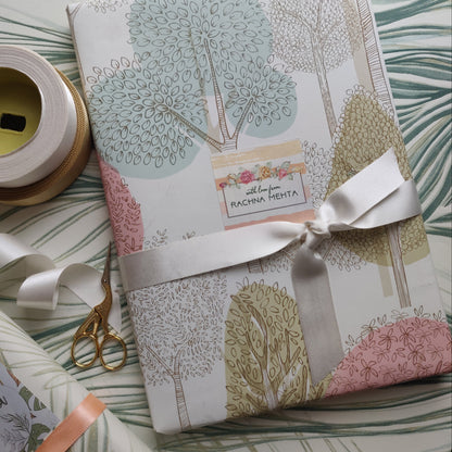 A wrapped gift box with a pastel tree-patterned paper wrap and a white satin ribbon bow, styled on a matching tropical leaf-patterned surface with gift ribbon spools, scissors, and a small card reading 'with love from PACHRA MENTHA'.