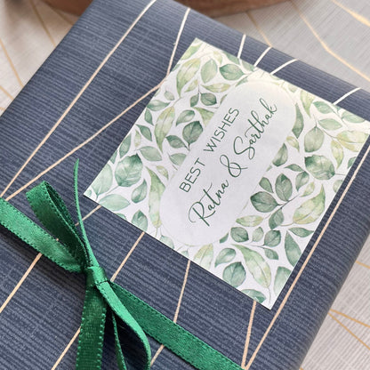 A wrapped gift box with a dark gray and white striped pattern, tied with a green ribbon and featuring a decorative watercolor leaf label that reads 'Best Wishes Relax & Smile'.