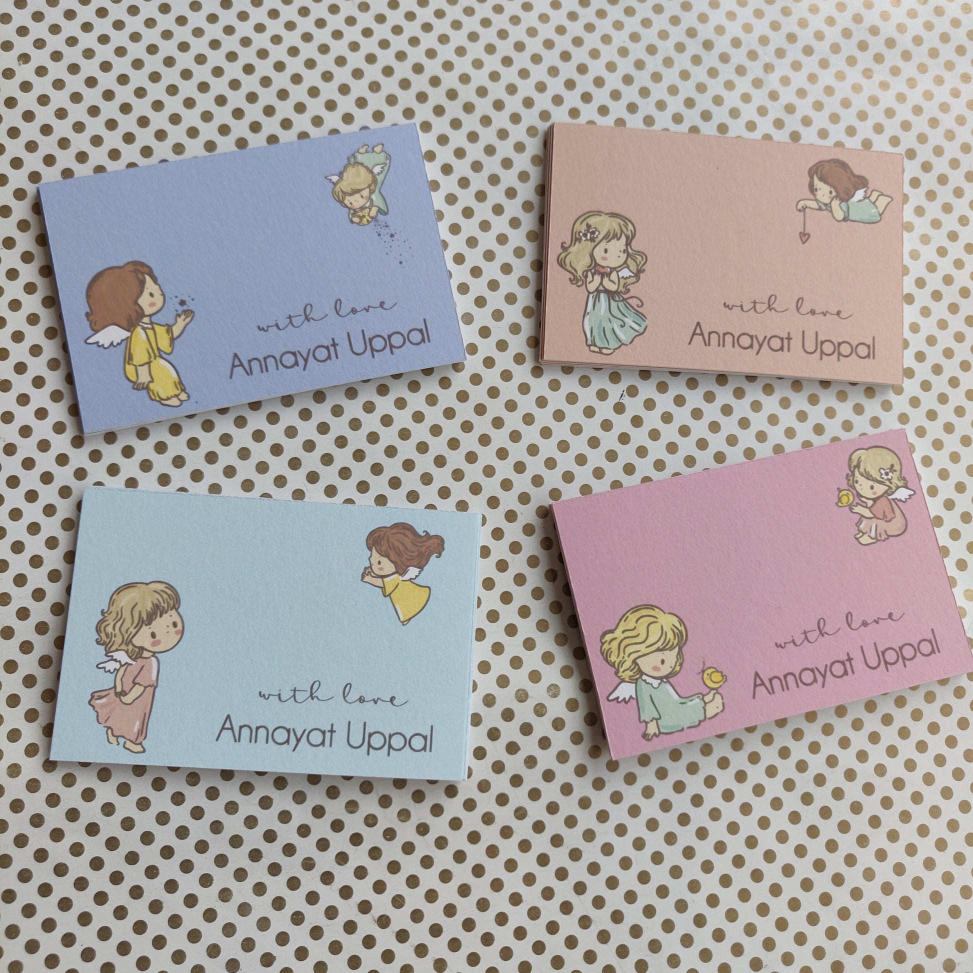 Four small rectangular cards arranged on a beige surface with a brown polka-dot pattern. Each card has a pastel background in blue, pink, or light blue, with a cute illustration of a girl and the text 'with love Annayat Uppal'.