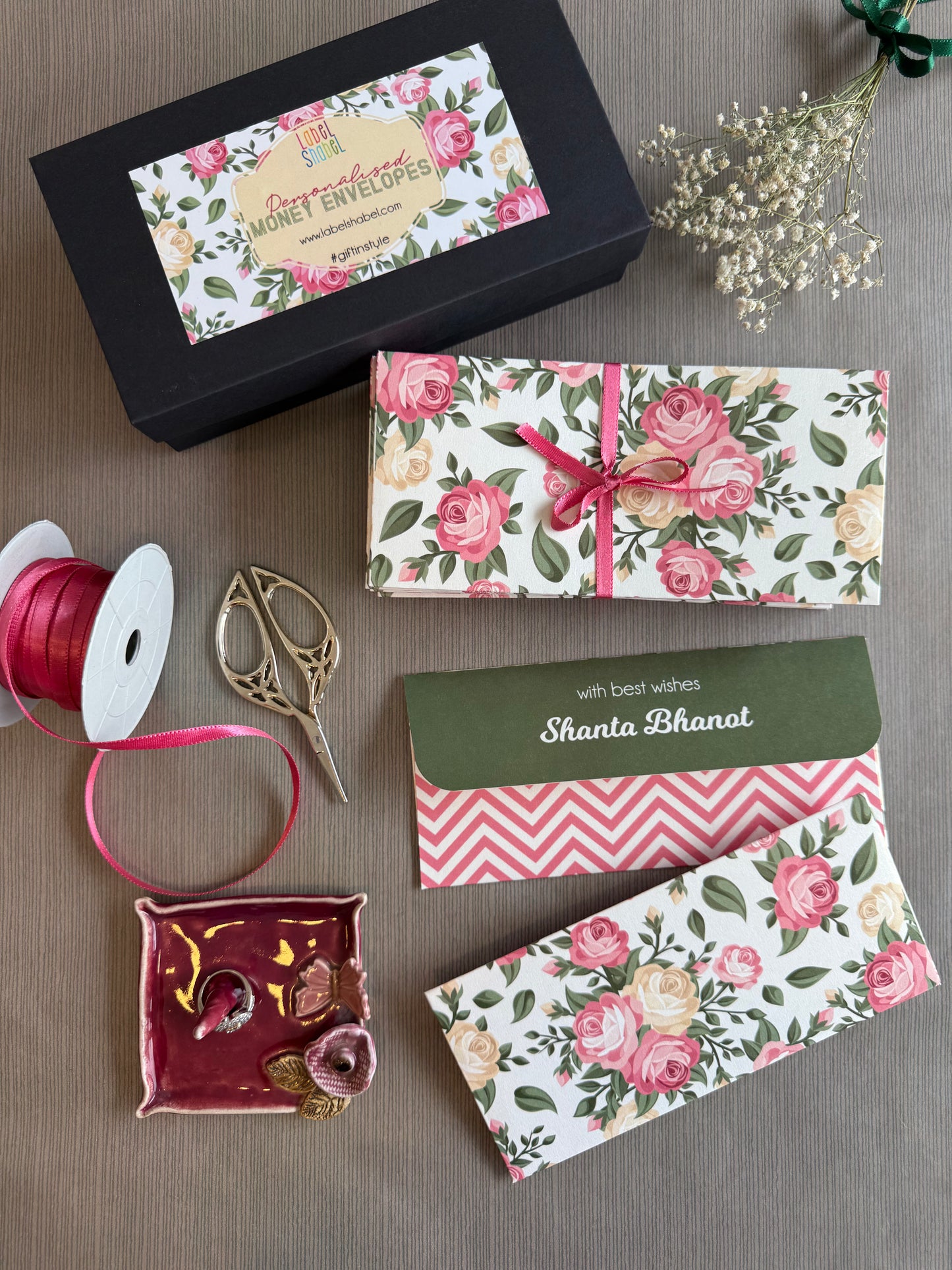 A styled flat-lay of floral honey envelopes and gift packaging on a neutral fabric background, including a black branded box, wrapped envelopes with pink roses, ribbon, scissors, and decorative elements.