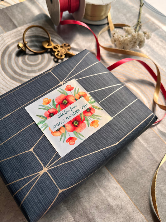 A wrapped gift box featuring a white floral label that reads 'Avalin's Made with Love' and decorated with red and orange flowers. Styled on a gray patterned fabric with ribbons and decorative props.