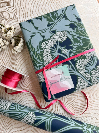A wrapped gift box and a roll of matching patterned wrapping paper on a beige wavy-textured surface, tied with a pink ribbon and a small card. The wrapping paper has a botanical design in navy, sage green, and white, with a pink ribbon accent.