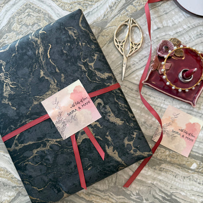 A wrapped gift box featuring a black and gold marble pattern, tied with a dusty red ribbon and adorned with a pink watercolor tag that reads 'Happy Valentine's Day'. The box is styled on a gray marble surface alongside decorative scissors and a small maroon dish.