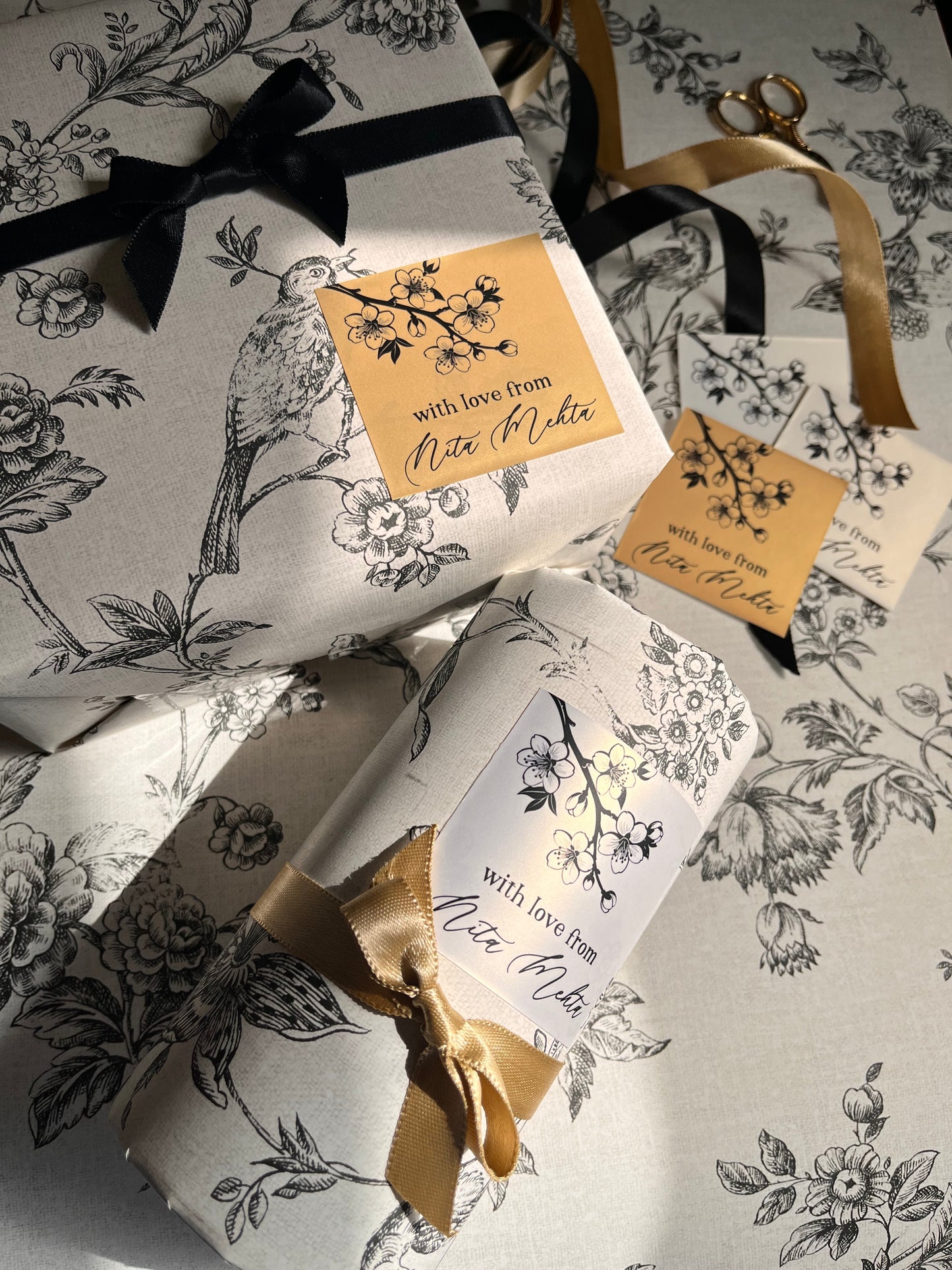Three gift boxes wrapped in matching white paper with a black floral and bird toile de Jouy pattern, tied with black and gold ribbons and decorated with gold tags reading 'with love, Nicole'.