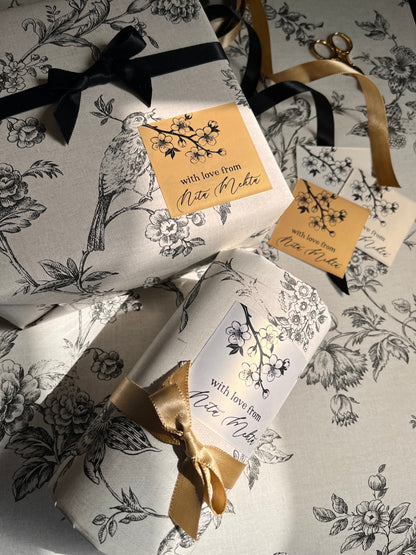 Three gift boxes wrapped in matching white paper with a black floral and bird toile de Jouy pattern, tied with black and gold ribbons and decorated with gold tags reading 'with love, Nicole'.