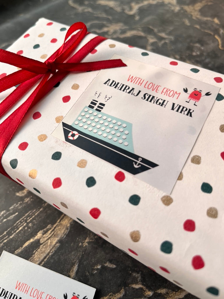 A gift box wrapped in white paper with red, green, and gold polka dots and tied with a red ribbon, featuring a printed card that reads 'WITH LOVE FROM ADHIRAJ SINGH VIRK' with a small boat illustration.