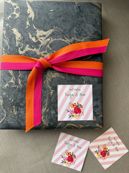 A wrapped gift box with dark gray and gold marble-patterned paper, tied with a bright orange and hot pink ribbon, on a gray wood surface. A square pink-and-white striped card with a floral illustration and the text 'best wishes Teigo & Nora' is attached to the box, with two matching cards placed in front.