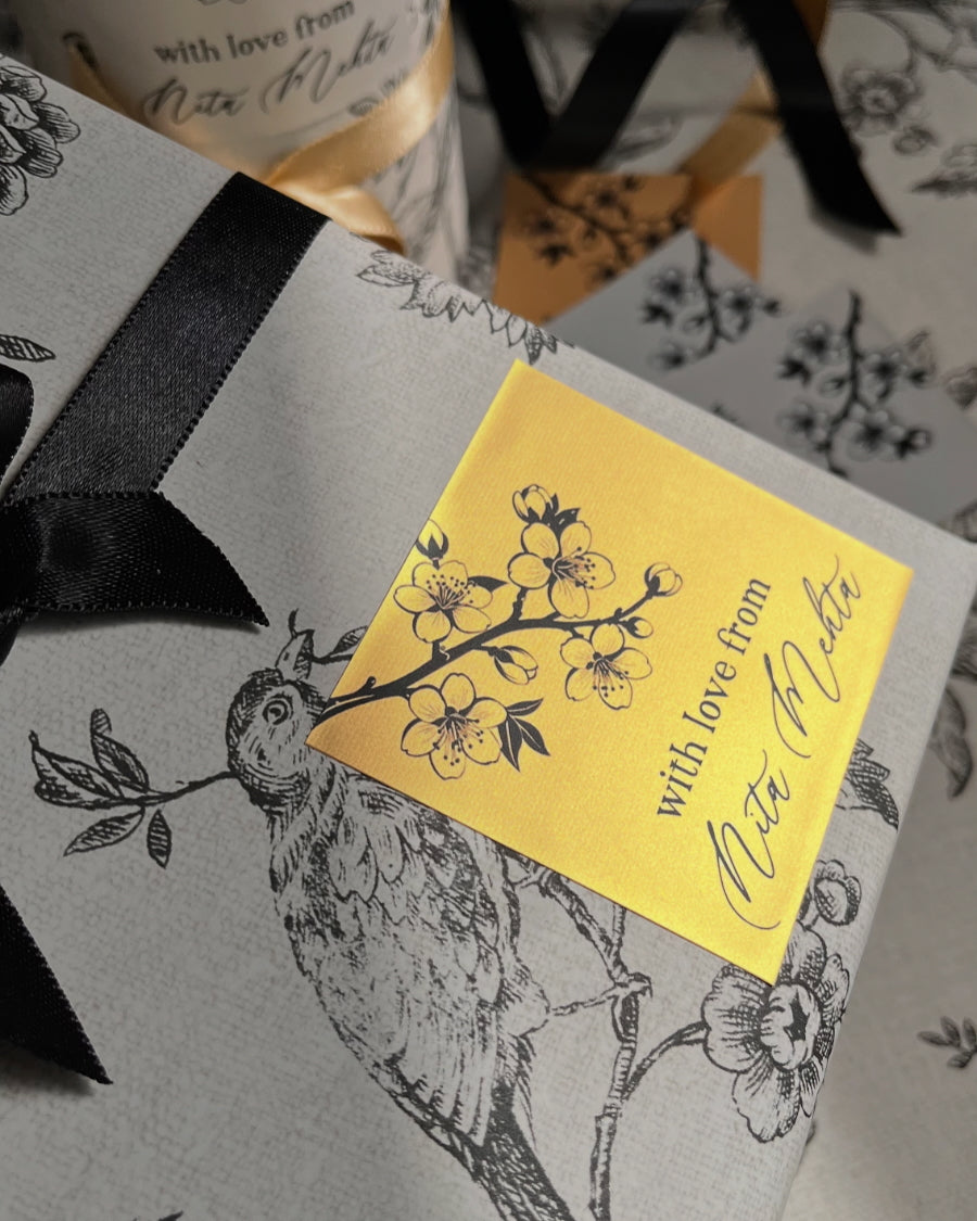 A close-up of a gift box or package with a light gray floral and bird print, tied with a black ribbon and topped with a yellow square card that reads 'with love from Rita Miller' with floral illustrations.