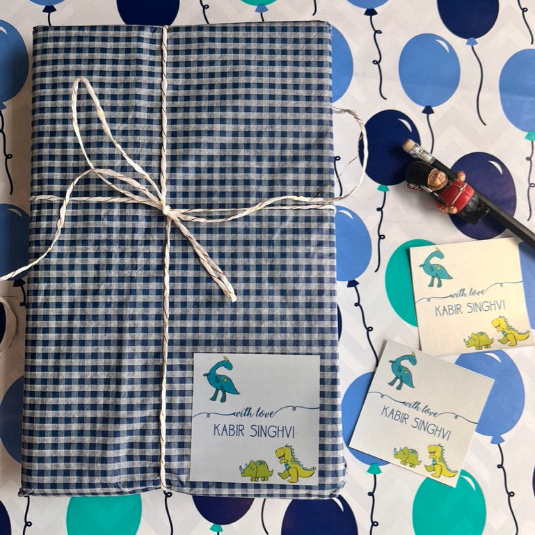 A gift-wrapped book or journal with a blue and white gingham pattern, tied with white string and featuring a personalized label that reads 'with love KABIR SINGHVI' with cartoon dinosaur illustrations. It is displayed on a white background patterned with blue and teal balloons, alongside two matching personalized cards and a small toy.