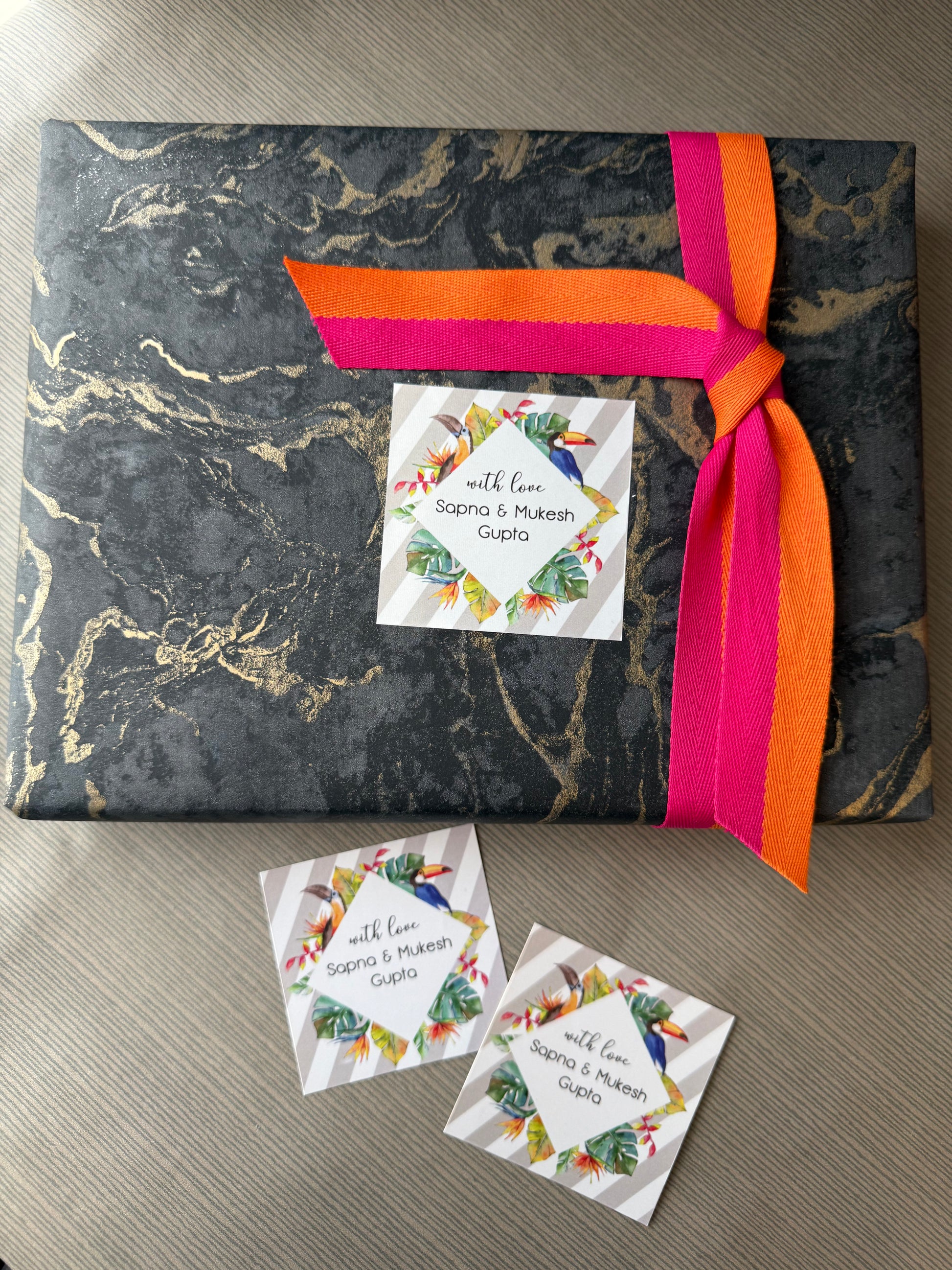A square gift box wrapped in black and gold marble-patterned paper, tied with a bright pink and orange ribbon, and decorated with a custom printed card. Two matching cards are placed on the wooden surface below.