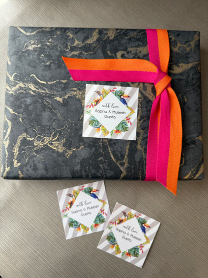 A square gift box wrapped in black and gold marble-patterned paper, tied with a bright pink and orange ribbon, and decorated with a custom printed card. Two matching cards are placed on the wooden surface below.